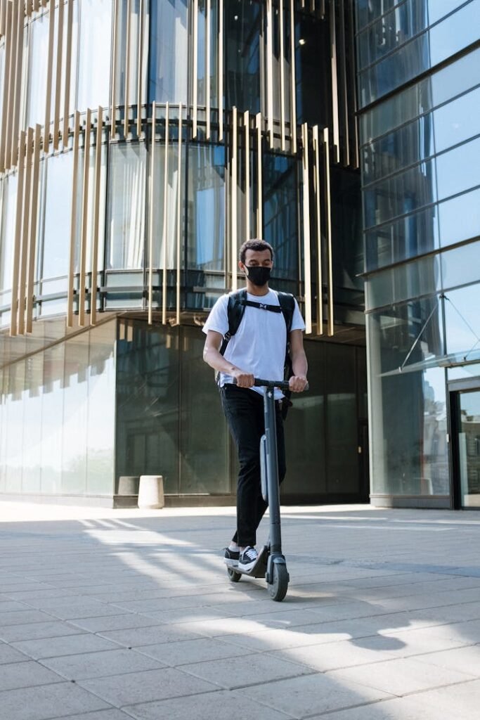 Man with backpack riding an electric scooter near a modern glass building in the city.